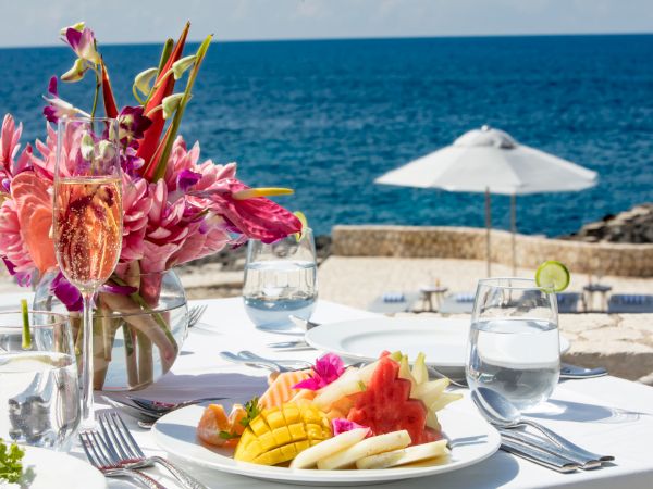 A seaside table set for a meal with tropical flowers, a fruit plate, water glasses, utensils, and a white umbrella over a sunny beach.