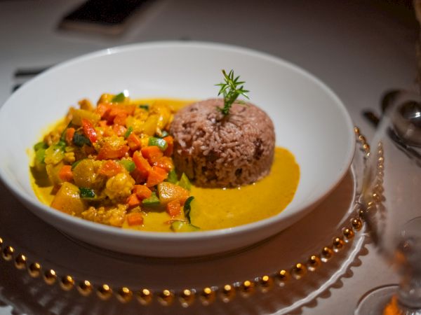 A plated dish with a colorful vegetable curry beside a mound of purple rice, all on a white plate with a yellow sauce drizzle.