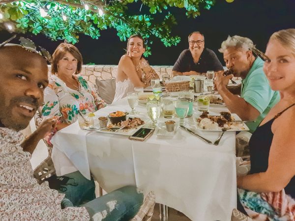 A group of six friends dining outdoors at a round table with white linens, enjoying drinks and desserts under string lights, smiling at the camera.