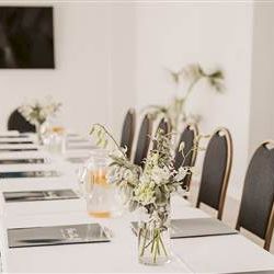 A long table setup for a meeting or event with white linens, small floral centerpieces, place settings, and black chairs along both sides.