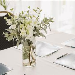 A vase with white flowers and green leaves sits on a white table, flanked by menus and a notebook in a bright, elegant dining setup.