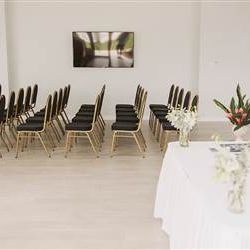 A small event room with rows of black chairs, a long table at the front draped in white, floral arrangements, and a TV on the wall.
