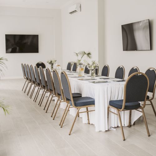 A long conference room with a U-shaped table setup, blue chairs, white tablecloths, a flat-screen on the wall, and plants by the windows.