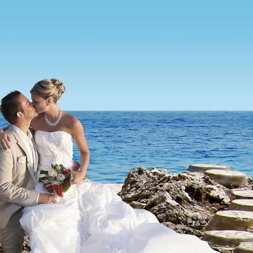 A newlywed couple sits by the sea, the groom in a beige suit and the bride in a white gown, sharing a kiss on rocky steps beside blue waters.