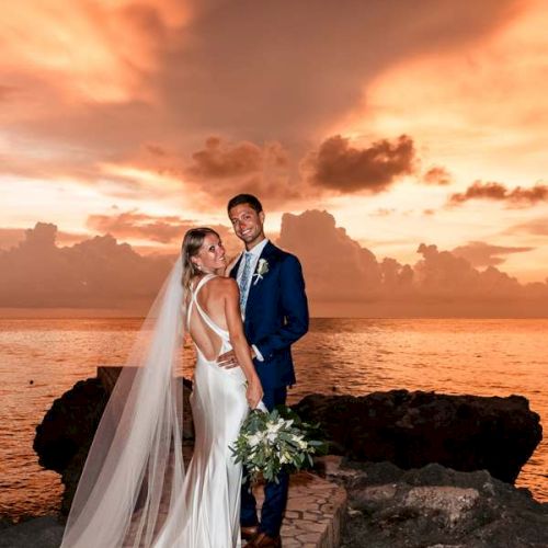 A newlywed couple stands on a rocky jetty at sunset, the bride in a white gown with a long veil and bouquet, the groom in a navy suit smiling beside her.