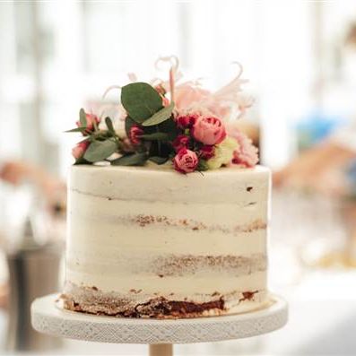 A simple, frosted cake with light beige buttercream and a small floral arrangement on top; blurred kitchen staff in the background, ending with a period.
