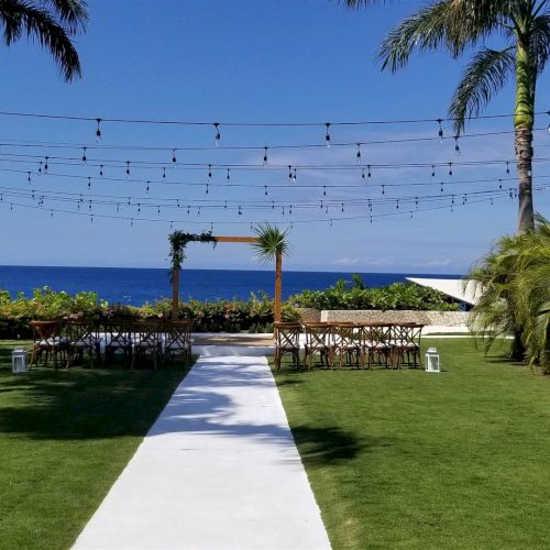A tropical resort scene with a stone path leading through lush green lawns to a blue ocean, palm trees lining the way, sunny skies.