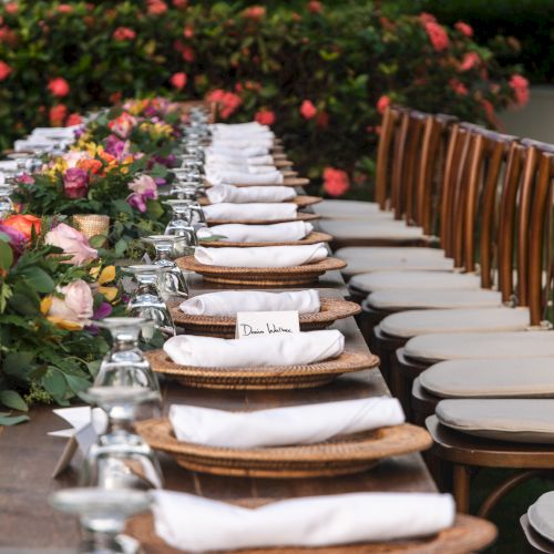 A long outdoor table setup with white plates, napkins, and glassware ready for a meal, surrounded by flower arrangements and greenery at a garden party.