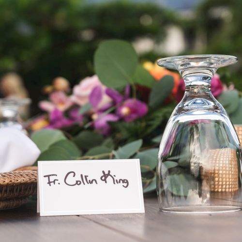 A table set for a formal event with a place card labeled &ldquo;Fr. Colin King,&rdquo; a glass upside down, floral centerpieces, and elegant decor.