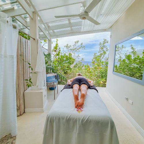 A person is lying face down on a massage table in a bright, airy spa room with a view of greenery and ocean beyond, relaxing.