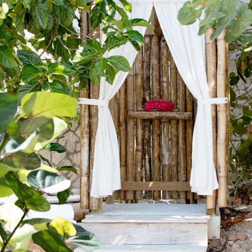 A small wooden cabana with white drapes tied back, a red flower centerpiece, surrounded by green foliage and a dirt/stone ground.
