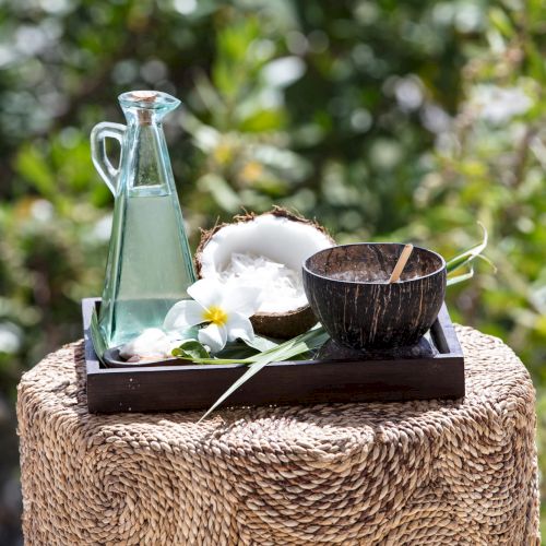 A glass bottle with a stopper, a small wooden bowl, a coconut half, a wooden tray, a flower, and a few glass cups on a woven stool outdoors, serene.