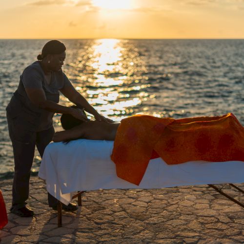 A person gives a massage on a table by the seaside at sunset, with a red cloth and small table nearby, relaxing scene by the ocean.