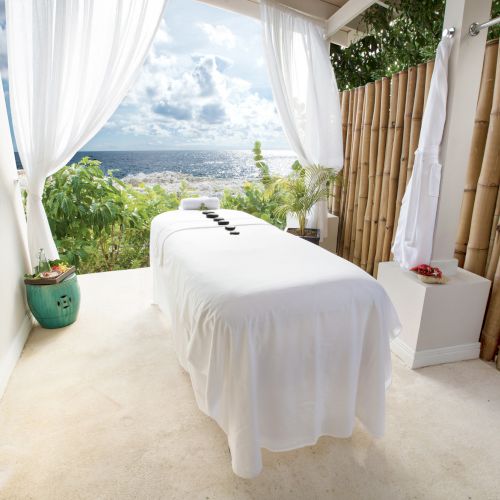 A luxurious outdoor massage table on a bright terrace with white curtains, greenery, and a scenic city view in the distance, serene and inviting.