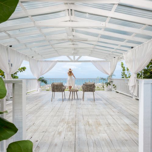 A person stands on a bright, white wooden deck under a white canopy, facing the ocean with chairs arranged around a table&mdash;tropical and serene.