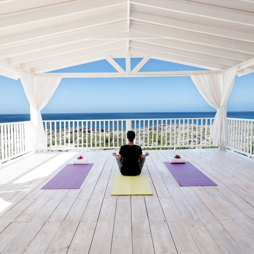 A person sits on a yellow yoga mat in a bright white open-air gazebo, facing the ocean, with two purple mats and a pink cup on either side, calm seaside serenity.