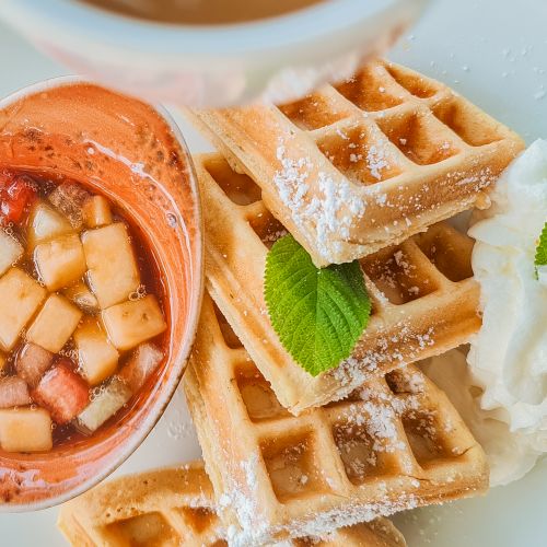 Delicious waffles stacked with powdered sugar, maple drizzle, cubed fruits in a side dip, and a dollop of whipped cream with a mint leaf, yum.