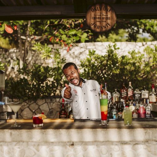 A smiling bartender in a white shirt behind a tropical outdoor bar, pointing at the camera, with colorful bottles and drinks on the counter.