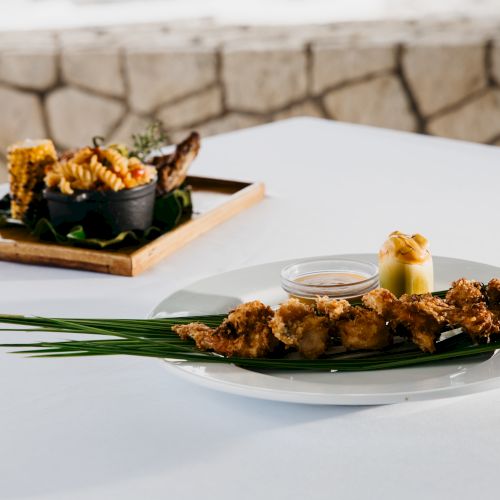 Sushi rolls and skewered fried bites on a white plate with greens, served on a patio table; a small dipping sauce sits beside.