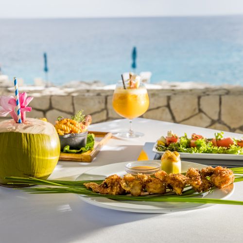 A sunny seaside meal with tropical drinks, grilled skewers, and fresh salads on a white-tabletop overlooking the ocean.