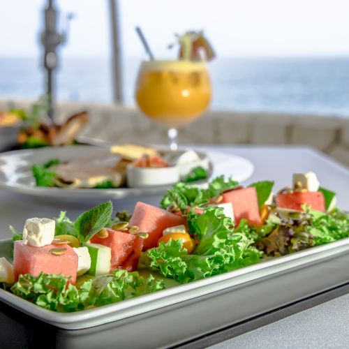 A vibrant plate of salad with watermelon and feta chunks on lettuce, garnished with herbs, on a seaside table; a drink and more food in the background.