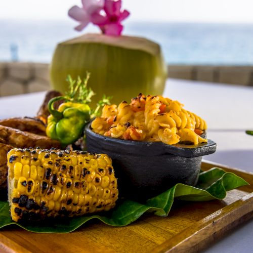Tropical beachside meal: grilled meat, corn on the cob, two bowls of golden rice with herbs, all served on a wooden board with a coconut drink.