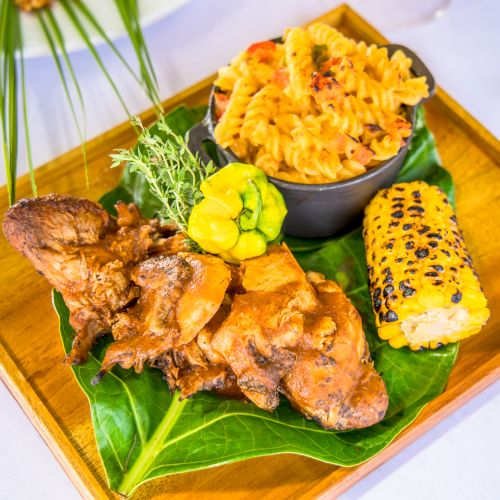 A plated meal featuring fried chicken pieces on a banana leaf, a side of corn with black sesame seeds, and a bowl of fried rice on a wooden tray.