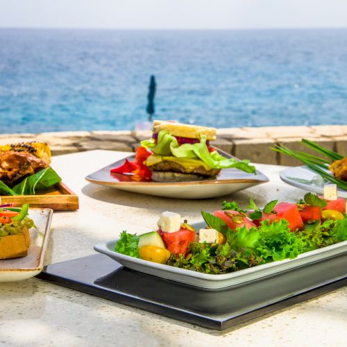 An oceanfront spread of colorful dishes on a white table: grilled sandwiches, fresh salads, and greens, with a blue sea backdrop.