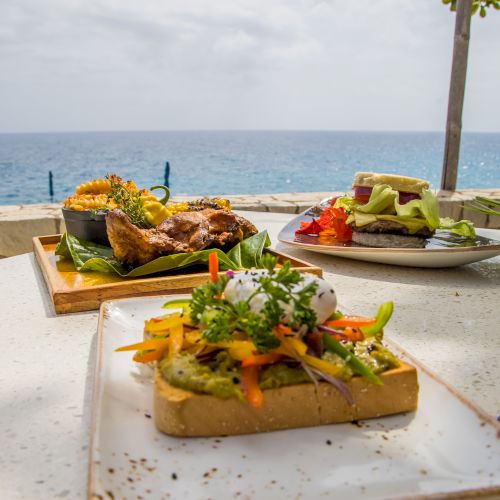 A seaside meal with three plated dishes on a light table, featuring toasted bread piles topped with veggies and herbs, and a bright ocean backdrop. (Note: ending punctuation should be period.)
