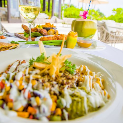 Delicious plated dish with creamy toppings, colorful veggies, and crispy garnish; glass of white wine and a sunny outdoor dining setup in the background.