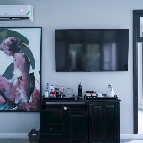 A stylish living space with a wall-mounted TV, a black cabinet topped with bottles, a large framed dog artwork, and a split air conditioner above.