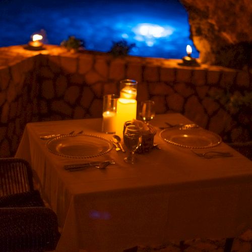 A candlelit romantic table setup on a terrace with a stone wall, dim lighting, two chairs, and a moonlit blue night sky.
