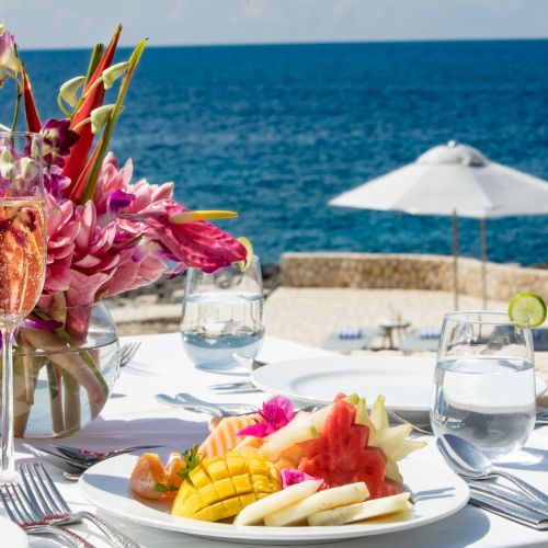A seaside table set for a meal with a colorful flower centerpiece, fruit plate, drinks, and a white umbrella by the ocean.