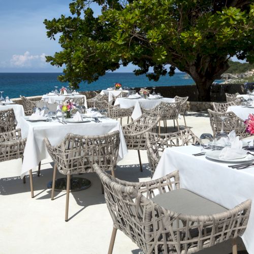 A beachside restaurant with white-tablecloth tables, wicker chairs, colorful flower centerpieces, and a view of the blue ocean under a leafy tree.