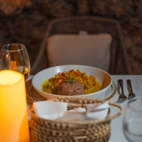 A candlelit table with a plated dish in a white bowl, surrounded by glassware, a bread basket, and a cozy, romantic dinner setting.