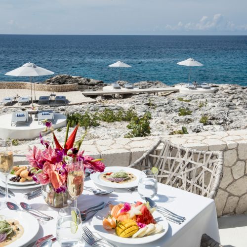 A seaside dining table set with white linens, flowers, and plates of food, stone terrace, sun umbrellas, and a blue ocean beyond the rocks. ending.