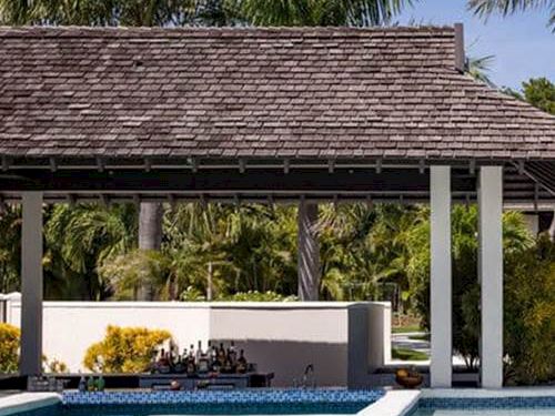 A resort-style pool with steps, a shaded gazebo, palm trees, and a sunny, tropical backdrop.