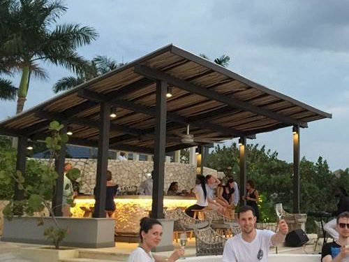 A few people sit and relax by a poolside bar with a wooden pavilion, palm trees, and a cloudy sky in the background.