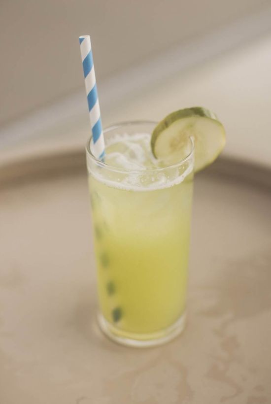 A tall glass of yellow lime drink with a striped blue straw and a lime wedge garnish, on a metal tray, looks refreshing.
