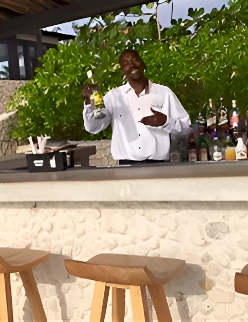 A bartender stands behind a white counter with bottles, chalkboard items, and wooden bar stools in an outdoor setting.
