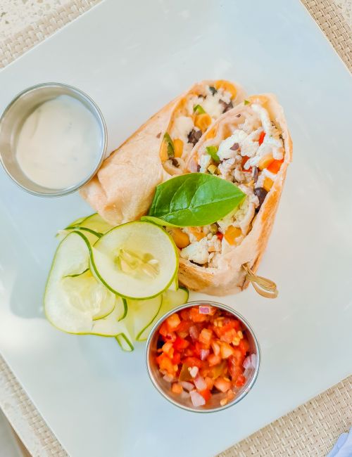 A veggie burrito on a plate with sliced cucumbers, a lime slice, a small bowl of salsa, and a dipping sauce on the side, delicious.