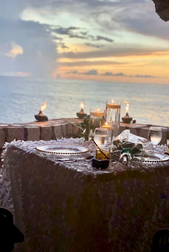 A romantic beachside dinner setup with candles, a lace tablecloth, and glowing sunset over the water, creating a serene, intimate scene.