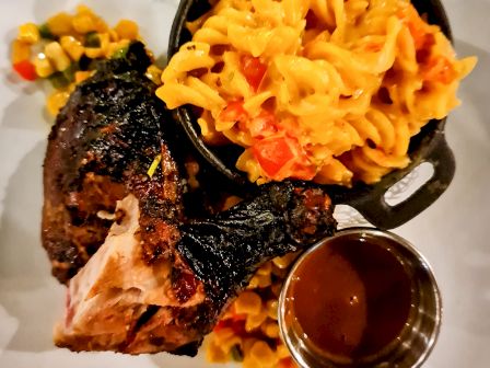 Smoked rib with charred crust, a side of cheesy macaroni, and a pool of dark barbecue sauce, plated over a bed of colorful corn and peppers.