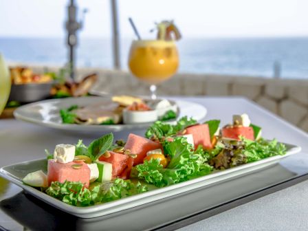 Fresh salad on a rectangular plate with greens, cubed watermelon or melon, feta, and herbs; seaside view with drinks in the background.