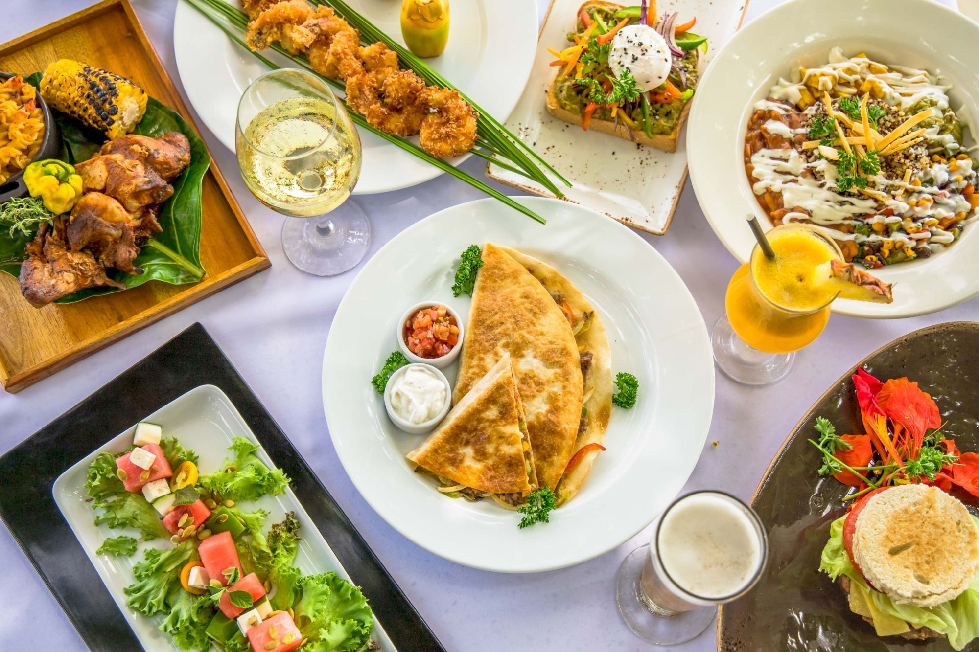 A colorful spread of dishes: quesadillas, tacos, greens, skewered bites, pasta, pancakes, and drinks on a white table.