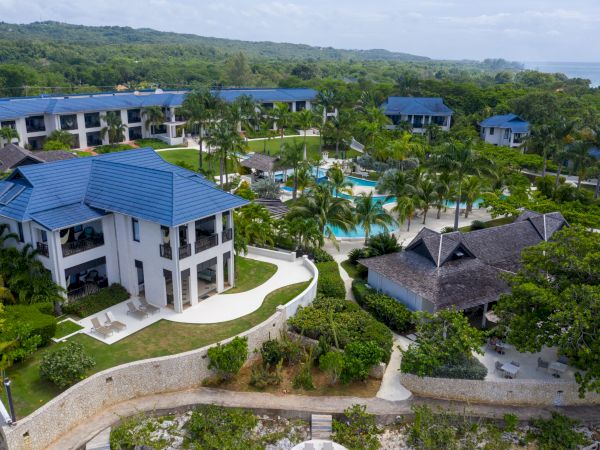 A resort with blue-roofed buildings, lush green gardens, a winding path, and a central pool area by the coast, tropical and serene.