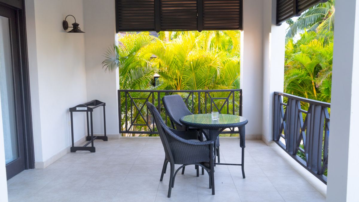 A balcony with a small table and two chairs, overlooking lush green palm trees; there&rsquo;s a railing, blinds, and a tiny stand against the wall, sunny outdoors.