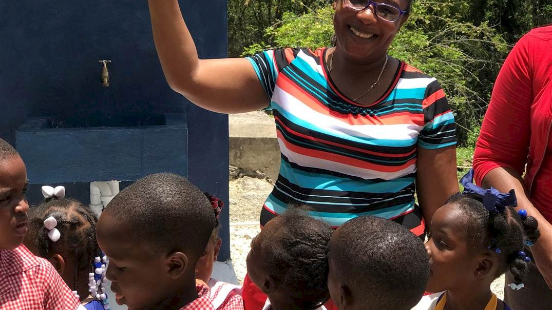 A smiling woman among children outside a colorful building, guiding them in a sunny outdoor setting, with everyone dressed in bright clothes.