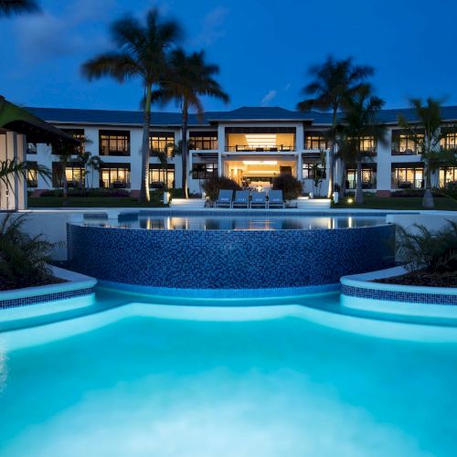A luxurious resort pool at dusk, illuminated turquoise water with palm-fringed surroundings and a grand hotel fa&ccedil;ade in the background.