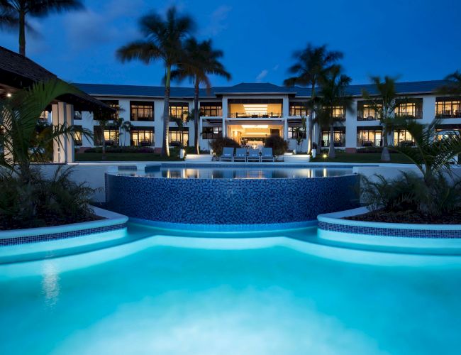 A luxurious resort pool at dusk, illuminated turquoise water with palm-fringed surroundings and a grand hotel fa&ccedil;ade in the background.